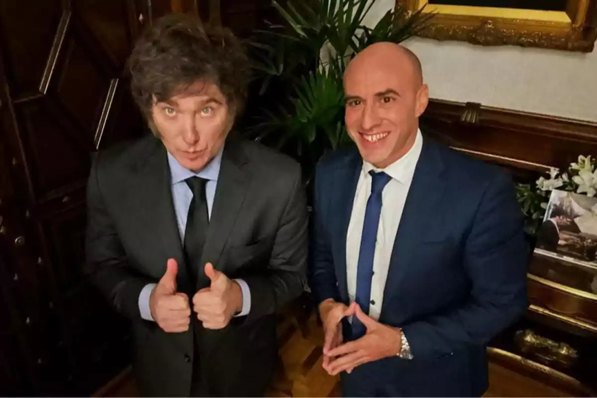 Two men dressed in suits pose smiling in an elegant setting with plants and a painting in the background.