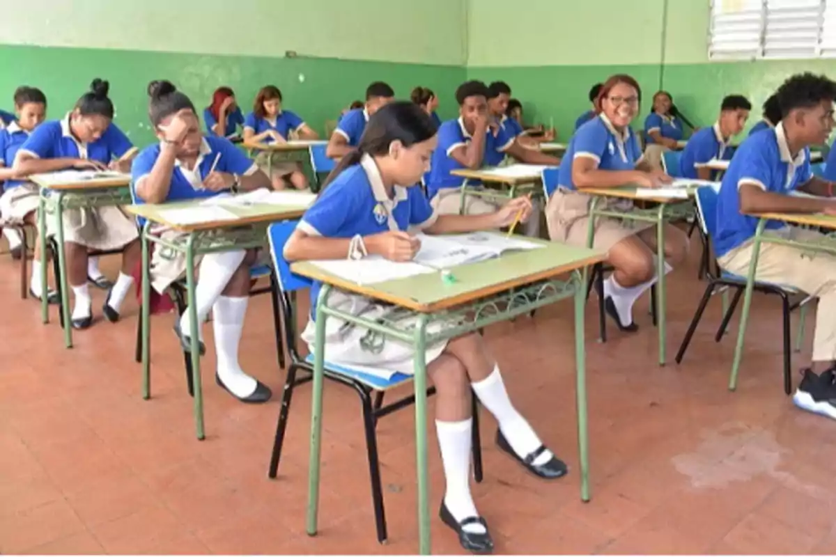 Estudiantes con uniforme escolar azul y beige sentados en pupitres dentro de un aula mientras realizan una actividad escrita