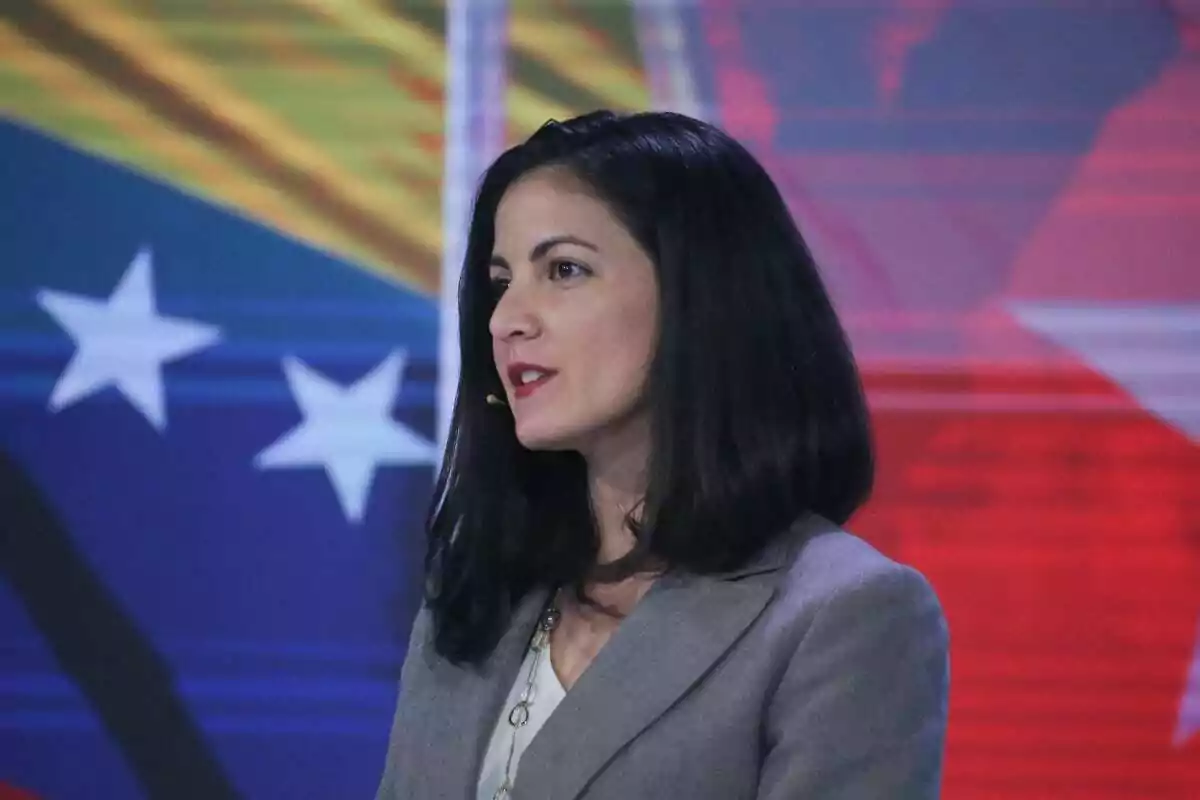 Mujer de cabello oscuro y liso usando saco gris y micrófono de diadema frente a un fondo colorido con estrellas blancas.