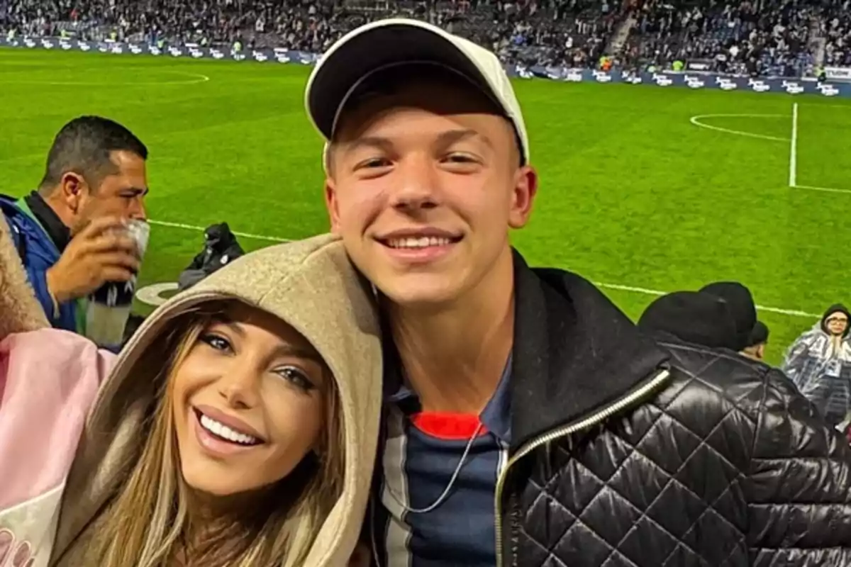 Two people smile as they pose for a photo in a soccer stadium full of spectators.