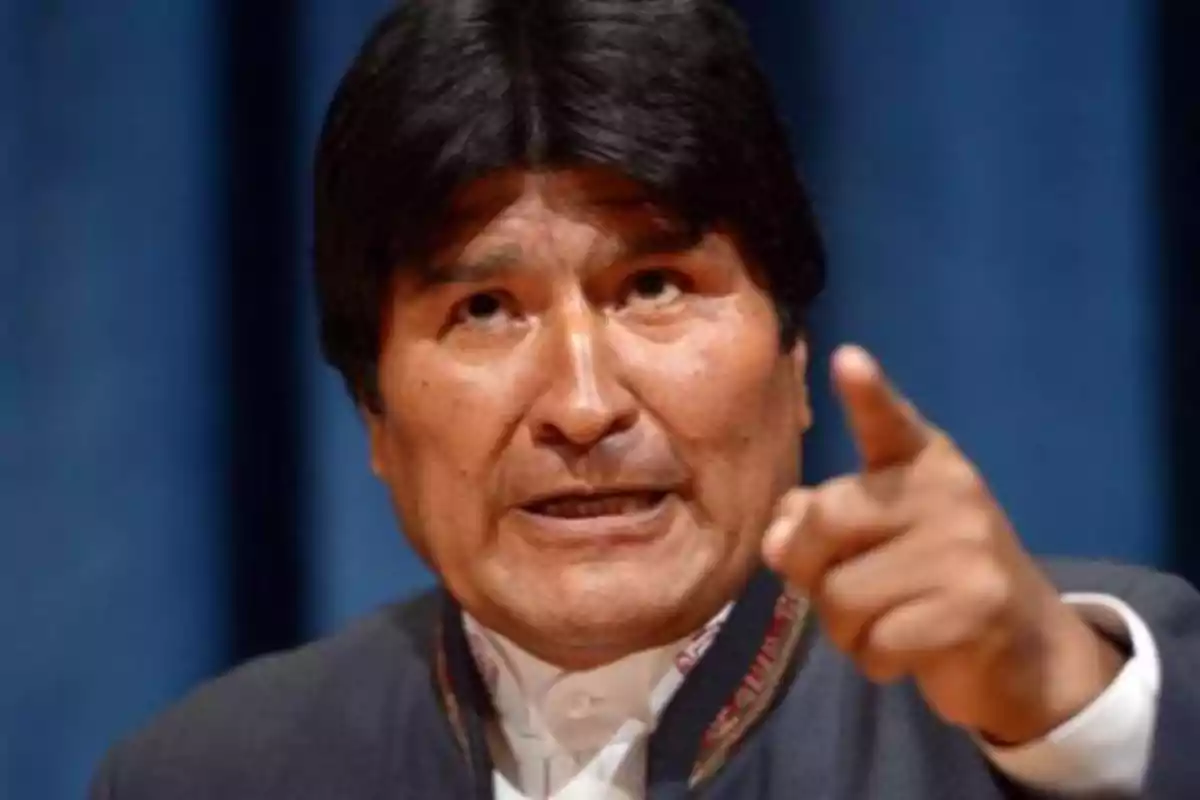 Hombre de cabello oscuro señalando con el dedo mientras habla con expresión seria frente a un fondo azul