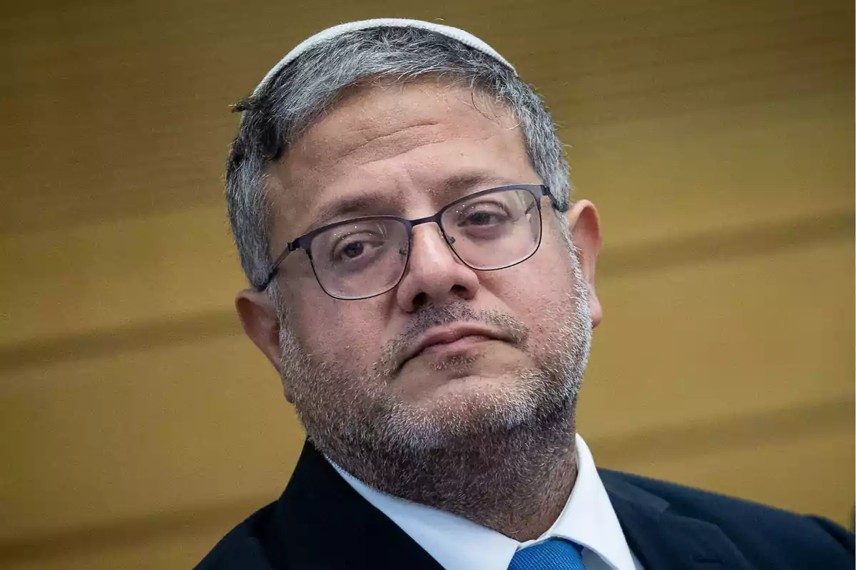 Middle-aged man with glasses, beard, and white kippah, wearing a suit and blue tie, looking straight ahead with a serious expression Middle-aged man with glasses, beard, and white kippah, wearing a suit and blue tie, looking straight ahead with a serious expression