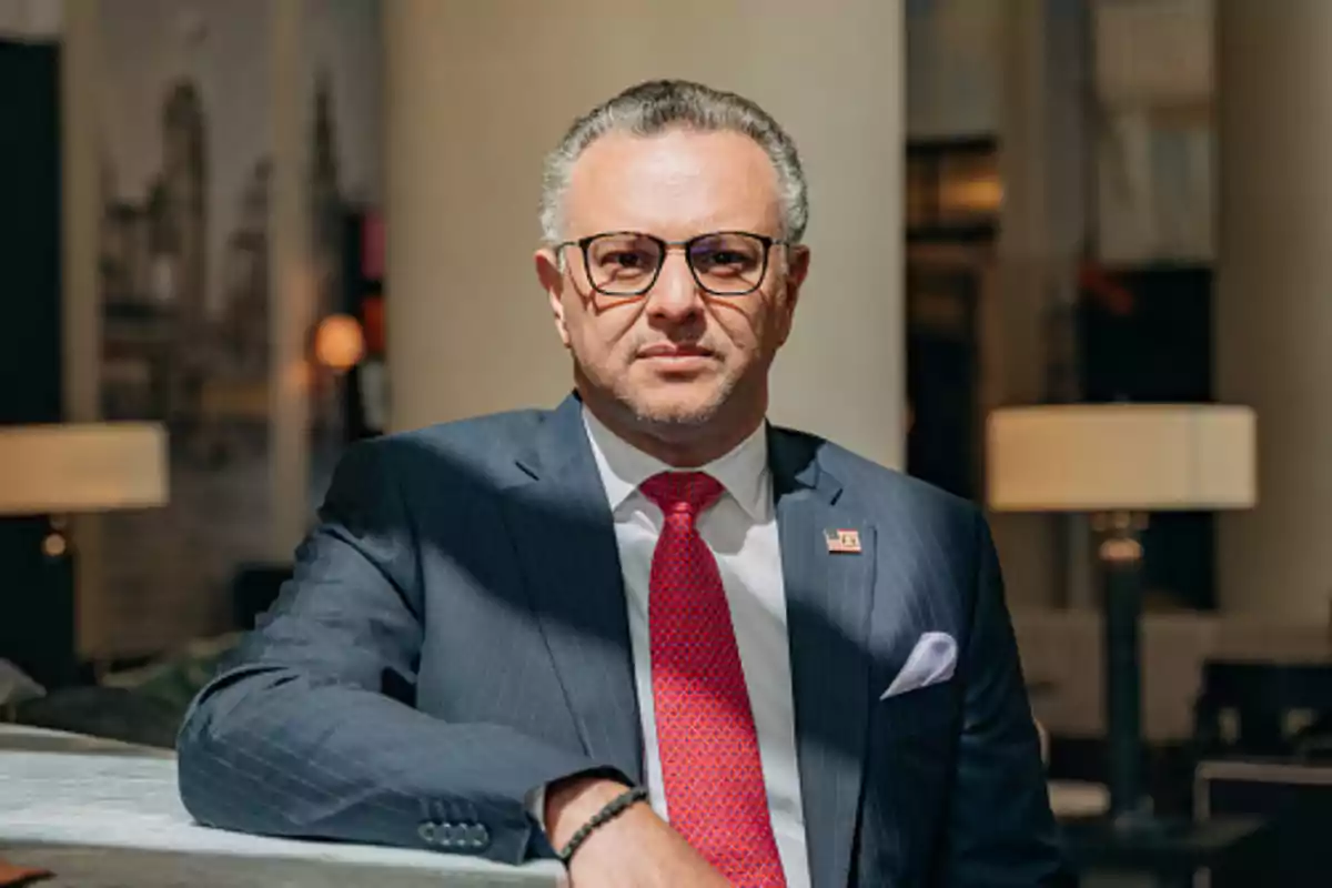 Hombre de mediana edad con gafas y traje azul oscuro con corbata roja sentado en un entorno elegante y bien iluminado Hombre de mediana edad con gafas y traje azul oscuro con corbata roja sentado en un entorno elegante y bien iluminado