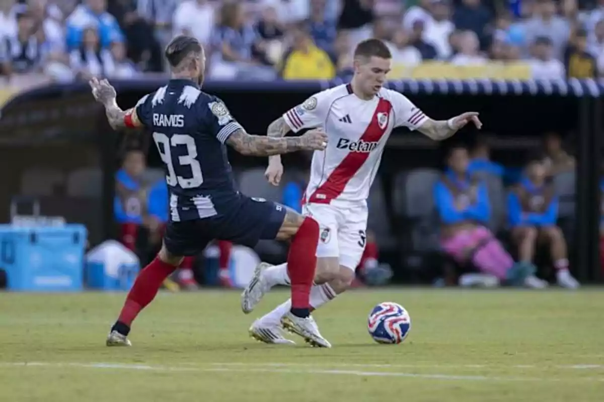 Two players compete for the ball during a match, one wearing a white uniform with a red stripe and the other in a dark blue uniform with the number 93 on his back, while the crowd watches from the stands. Two players compete for the ball during a match, one wearing a white uniform with a red stripe and the other in a dark blue uniform with the number 93 on his back, while the crowd watches from the stands.