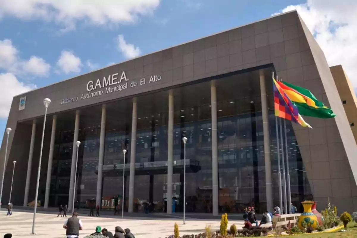 Fachada moderna del edificio del Gobierno Autónomo Municipal de El Alto con varias banderas ondeando y personas en la plaza frontal Fachada moderna del edificio del Gobierno Autónomo Municipal de El Alto con varias banderas ondeando y personas en la plaza frontal