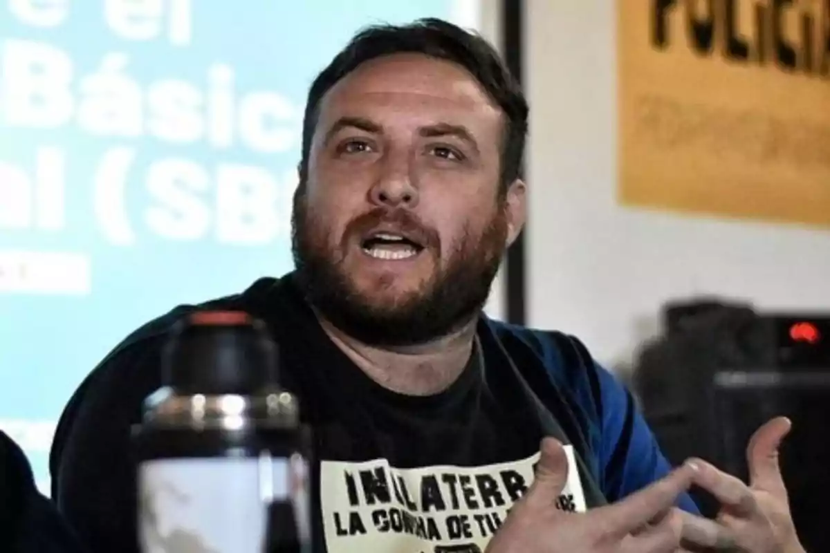 Bearded man speaking and gesturing with his hands in an indoor setting with posters and a blue board in the background Bearded man speaking and gesturing with his hands in an indoor setting with posters and a blue board in the background