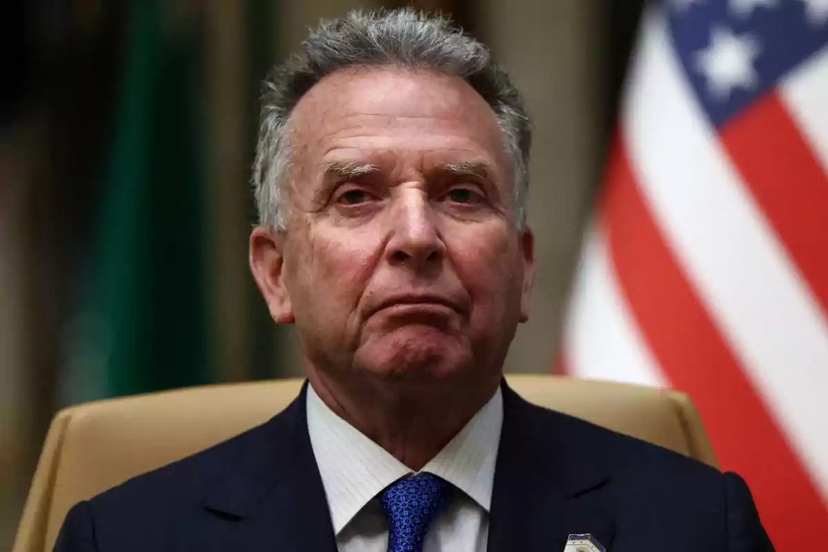 Hombre de cabello canoso con expresión seria vistiendo traje oscuro y corbata azul sentado frente a una bandera de Estados Unidos Hombre de cabello canoso con expresión seria vistiendo traje oscuro y corbata azul sentado frente a una bandera de Estados Unidos