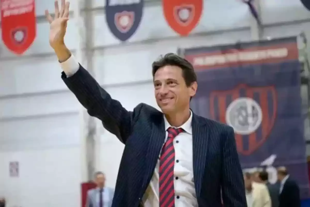 Man in a suit and tie smiles and waves, raising his hand in a sports stadium with pennants and a crest in the background.