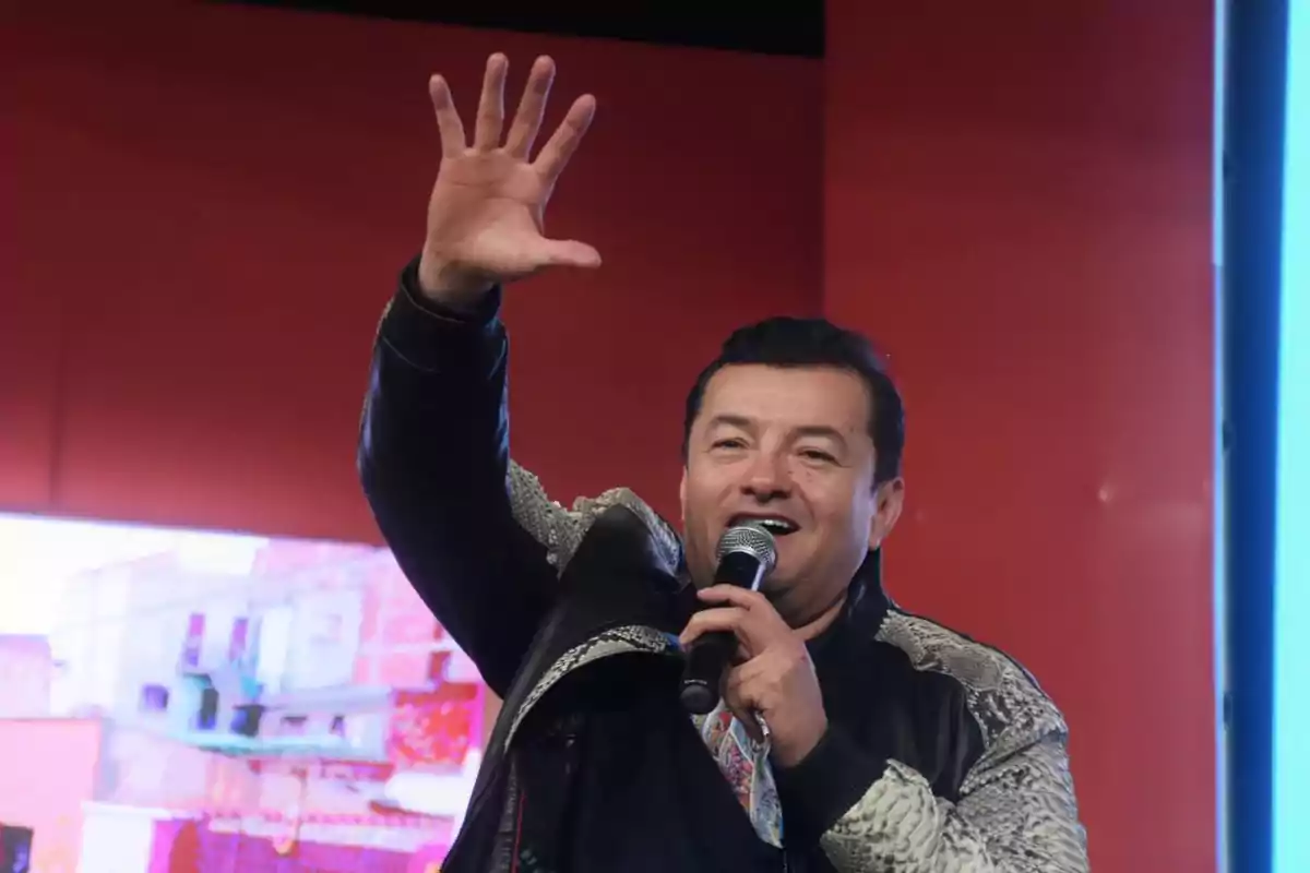 Hombre sonriente con micrófono en la mano derecha levantando la mano izquierda en señal de saludo sobre un fondo rojo Hombre sonriente con micrófono en la mano derecha levantando la mano izquierda en señal de saludo sobre un fondo rojo
