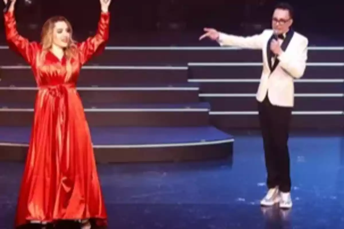 A woman in a red dress raises her arms while a man in a white jacket with a microphone points at her on a stage with stairs in the background. A woman in a red dress raises her arms while a man in a white jacket with a microphone points at her on a stage with stairs in the background.