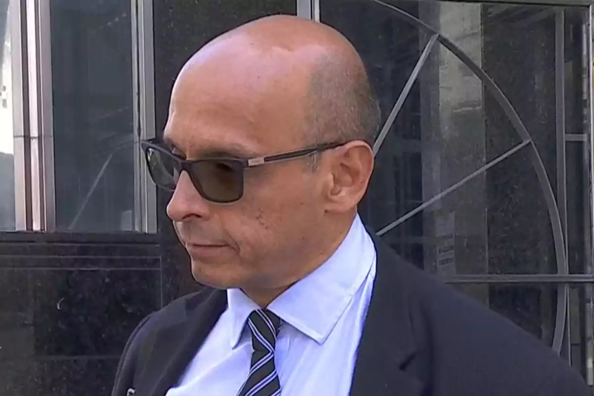 Hombre calvo con gafas oscuras y traje formal frente a un edificio de vidrio y metal Hombre calvo con gafas oscuras y traje formal frente a un edificio de vidrio y metal