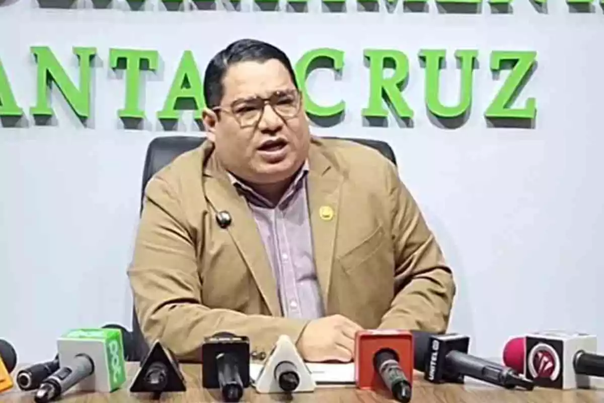 Man with glasses and a beige jacket speaking at a press conference in front of several microphones with green letters in the background