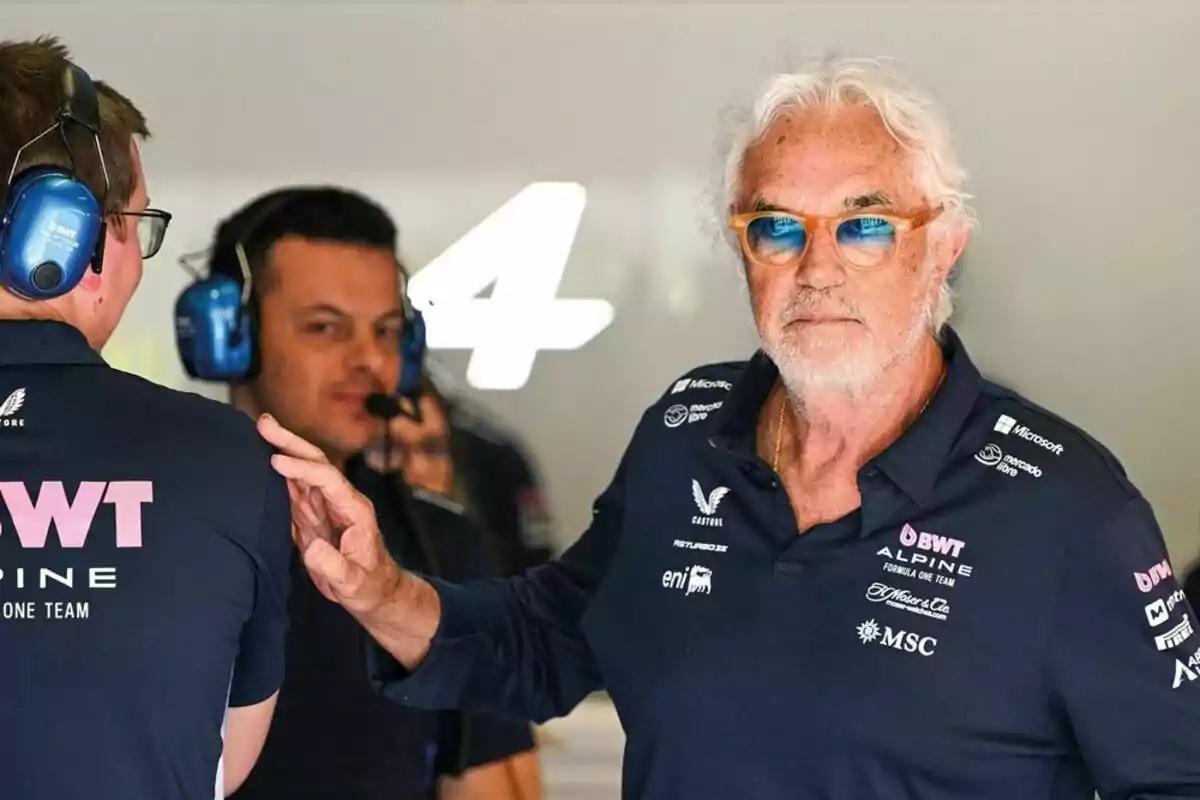 Gray-haired man with blue sunglasses wearing an Alpine team jersey in a Formula 1 garage next to two people with communication headsets Gray-haired man with blue sunglasses wearing an Alpine team jersey in a Formula 1 garage next to two people with communication headsets