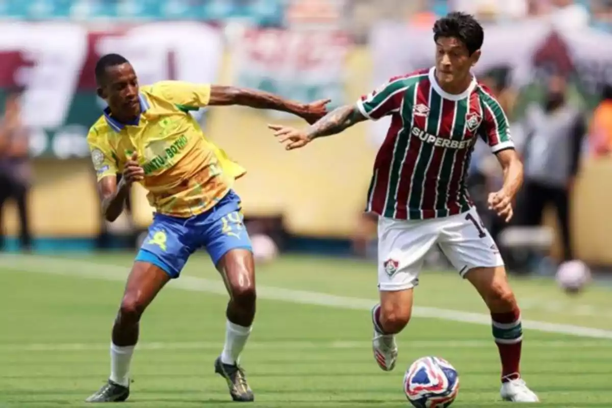 Two players compete for the ball during a match in a stadium, one wearing a yellow and blue uniform and the other wearing a green, red, and white striped uniform. Two players compete for the ball during a match in a stadium, one wearing a yellow and blue uniform and the other wearing a green, red, and white striped uniform.
