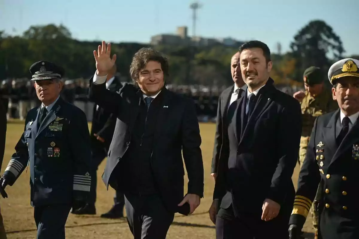 A man in a suit waves while walking accompanied by military officers at an outdoor event.