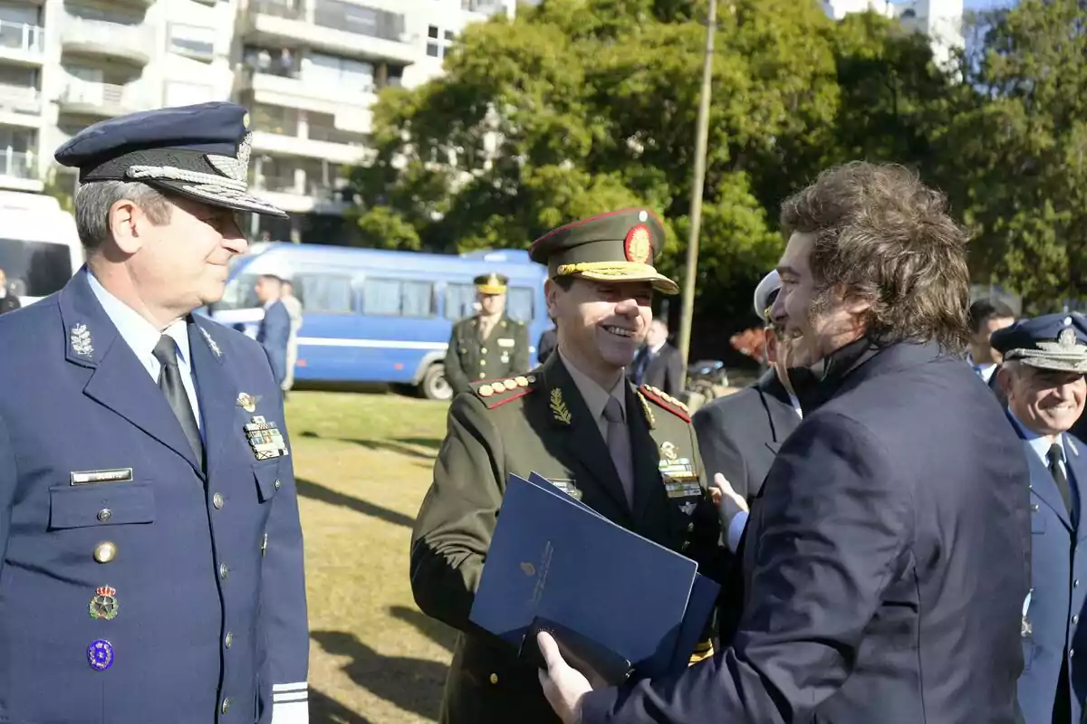 A group of men in military uniforms and suits talk and smile outdoors during an official event. A group of men in military uniforms and suits talk and smile outdoors during an official event.