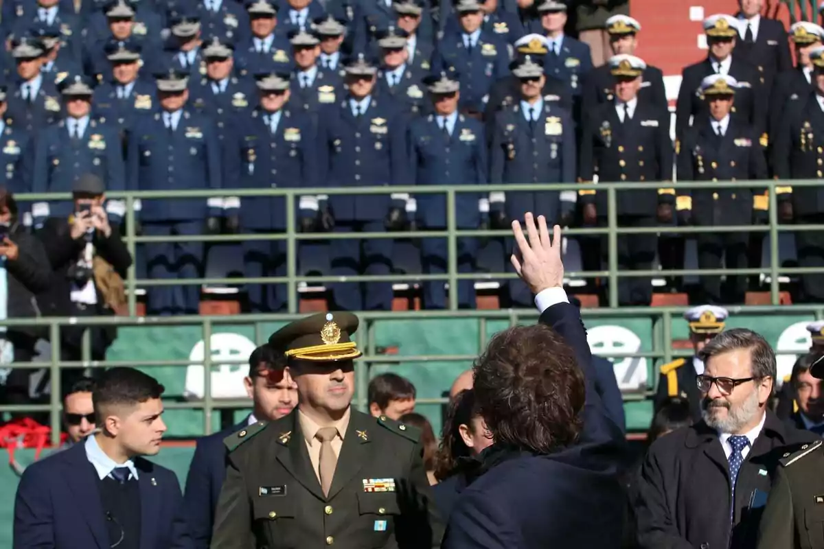 A man with his back turned raises his hand in front of a large group of uniformed soldiers standing on bleachers. A man with his back turned raises his hand in front of a large group of uniformed soldiers standing on bleachers.