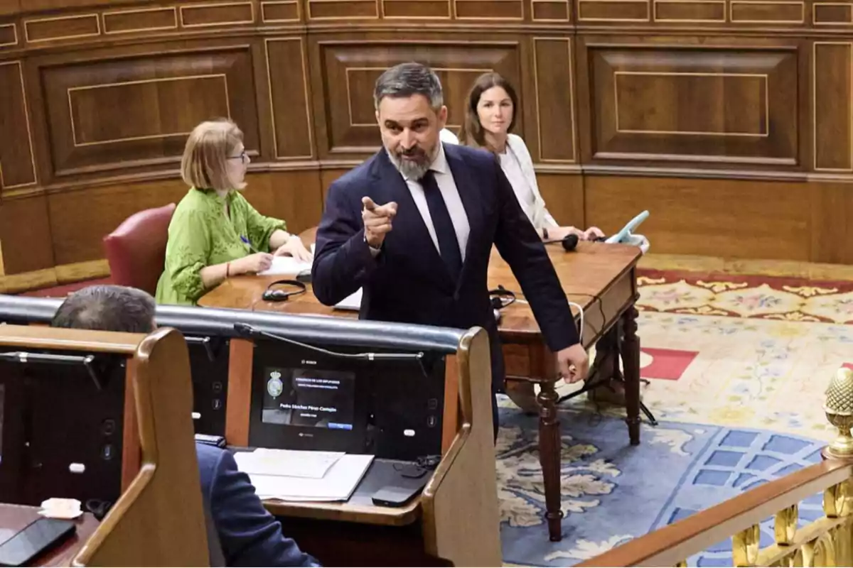 Un hombre de traje oscuro señala con el dedo mientras habla en una sala parlamentaria con dos mujeres sentadas al fondo y escritorios con pantallas en primer plano