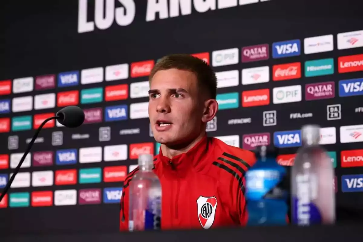 River Plate player wearing a red jacket speaking at a press conference with a sponsor backdrop and water bottles on the table