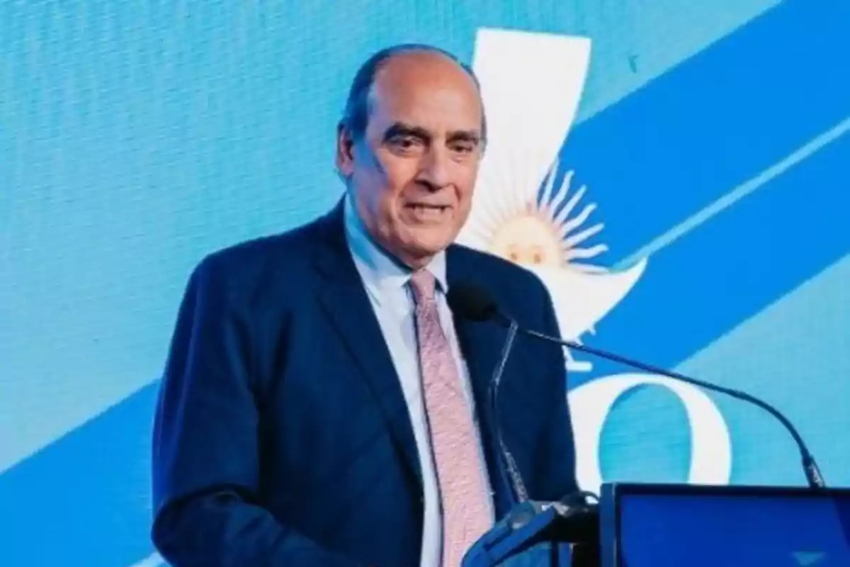 Hombre de traje hablando en un podio con fondo azul y un símbolo patrio detrás