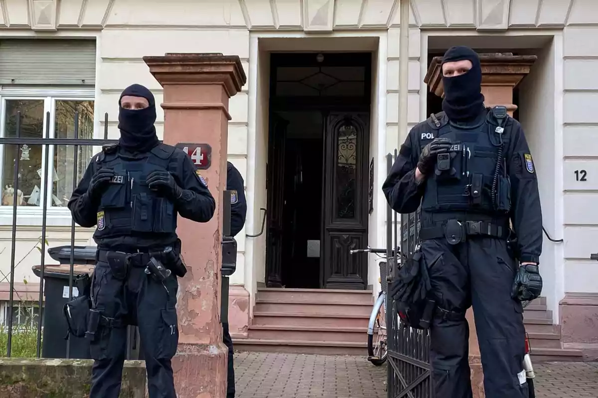 Dos policías con uniforme táctico y pasamontañas están de pie frente a la entrada de un edificio residencial Dos policías con uniforme táctico y pasamontañas están de pie frente a la entrada de un edificio residencial