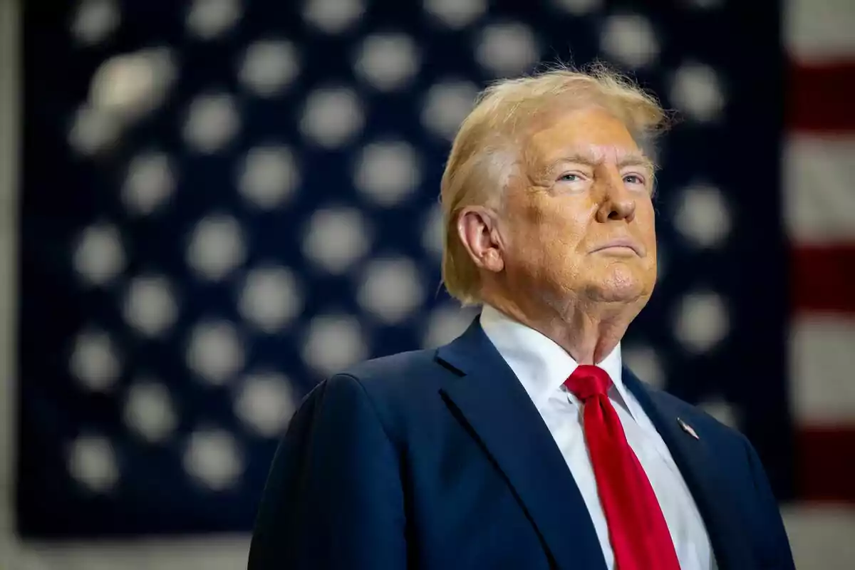 Blond-haired man in a blue suit and red tie in front of a blurred United States flag