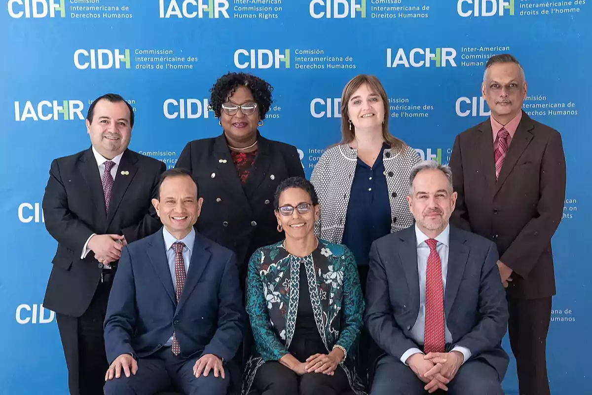 Seven people pose smiling in front of a blue background with logos of the Inter-American Commission on Human Rights. Seven people pose smiling in front of a blue background with logos of the Inter-American Commission on Human Rights.