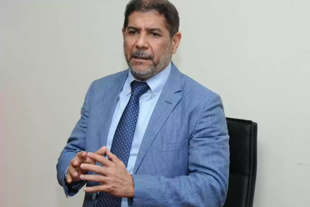 Hombre de mediana edad con barba y bigote, vestido con traje azul claro y corbata, sentado en una silla negra y gesticulando con las manos mientras habla.
