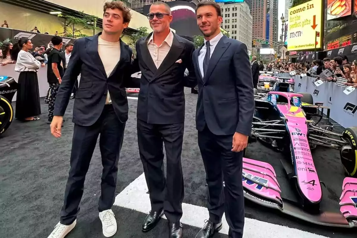 Tres hombres vestidos de traje posan juntos frente a un auto de Fórmula 1 color rosa en un evento al aire libre con público y edificios de fondo Tres hombres vestidos de traje posan juntos frente a un auto de Fórmula 1 color rosa en un evento al aire libre con público y edificios de fondo