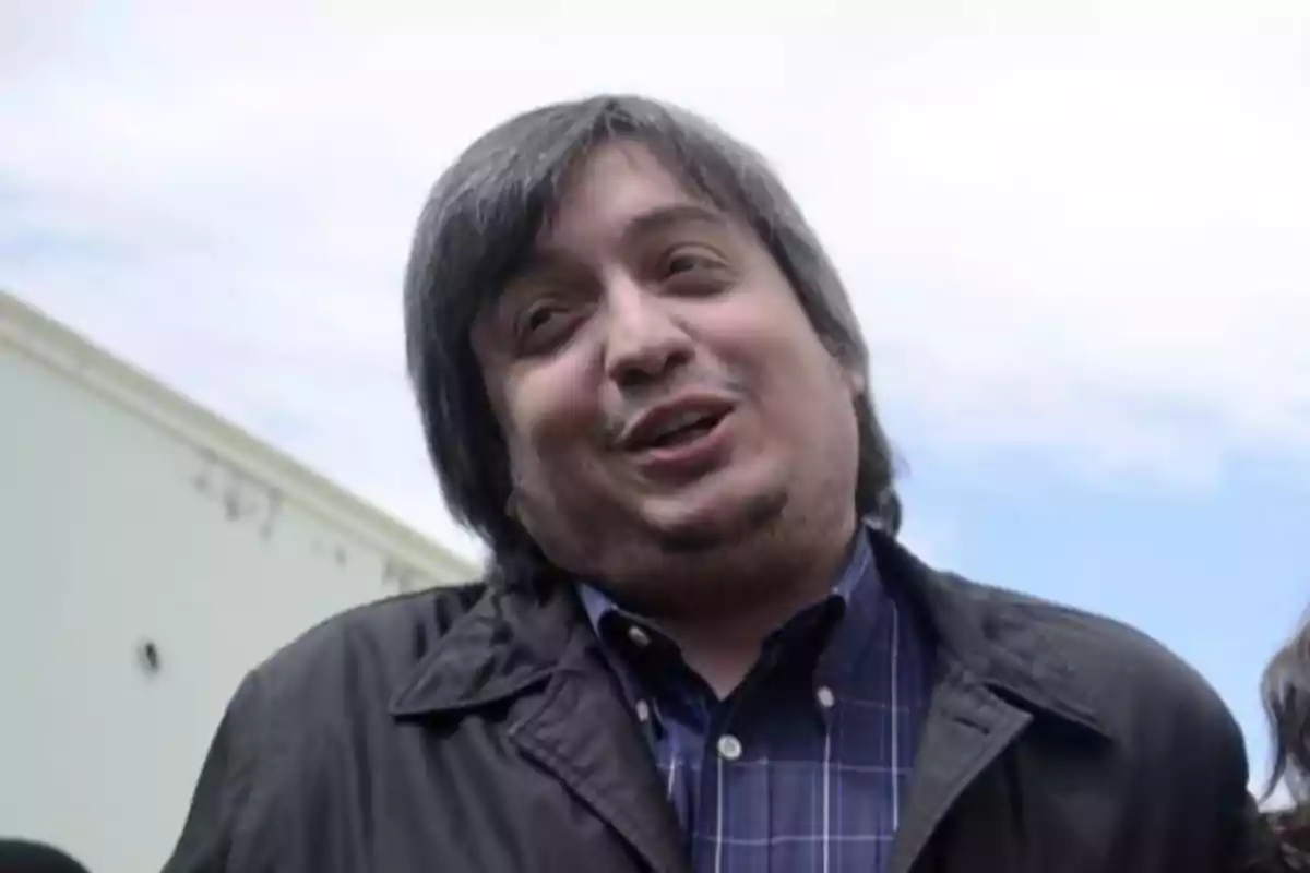 Hombre de cabello canoso y barba corta con expresión sonriente usando camisa a cuadros y chaqueta oscura al aire libre