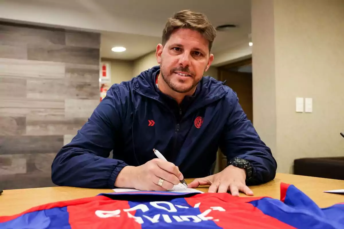 Man in a blue jacket signing a document on a table with a red and blue soccer jersey in the foreground Man in a blue jacket signing a document on a table with a red and blue soccer jersey in the foreground