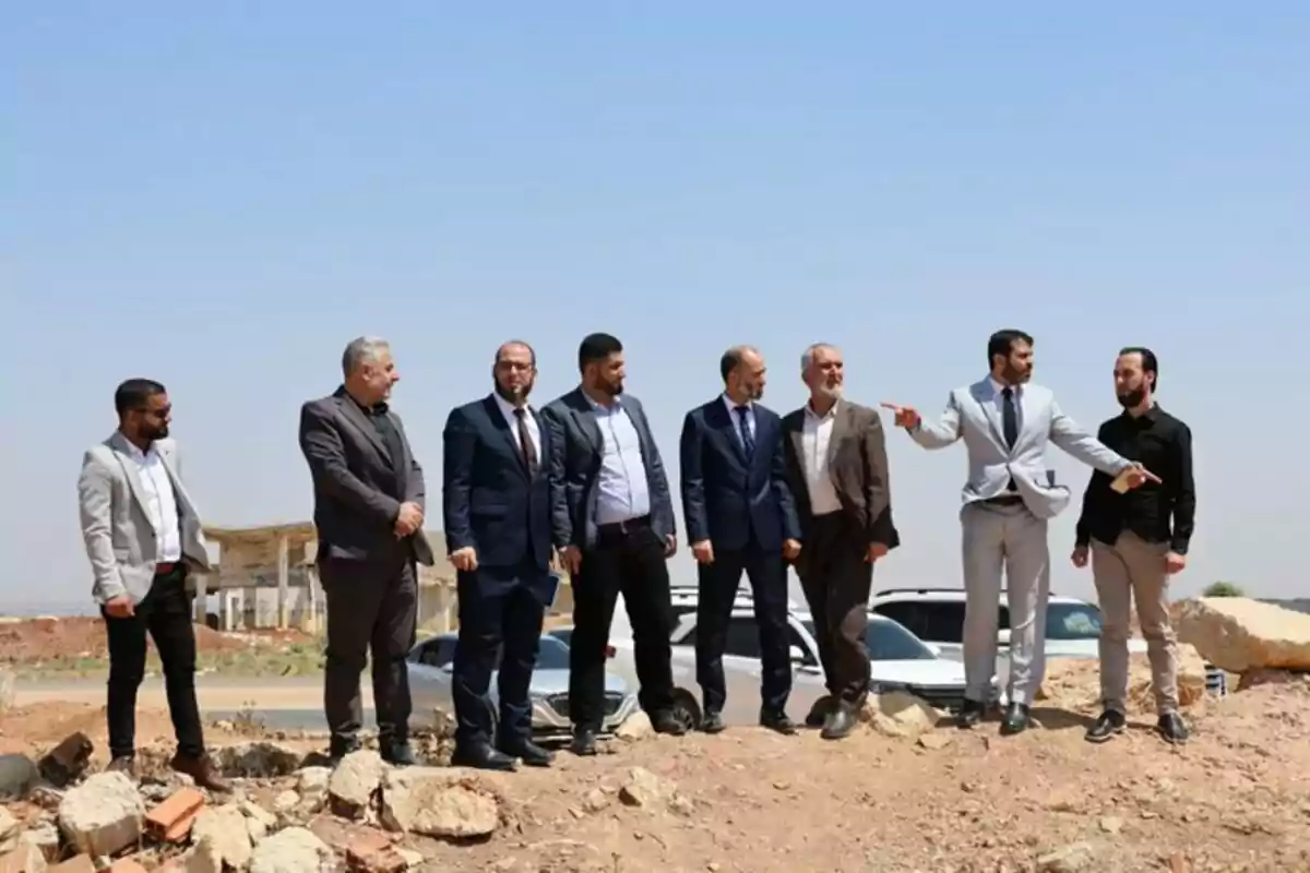 Ocho hombres vestidos de traje y ropa formal están de pie sobre un terreno árido con escombros mientras conversan y observan el lugar bajo un cielo despejado Ocho hombres vestidos de traje y ropa formal están de pie sobre un terreno árido con escombros mientras conversan y observan el lugar bajo un cielo despejado