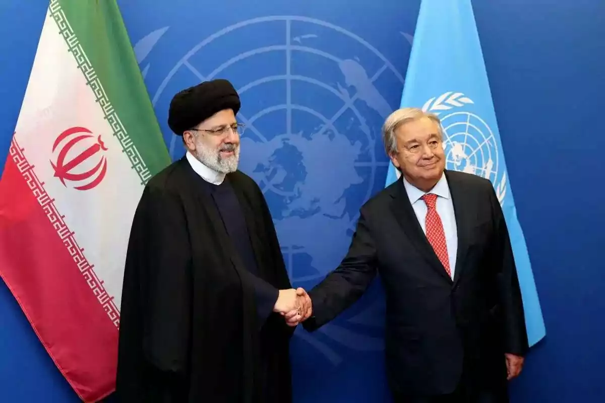 Two men in suits shake hands in front of the flags of Iran and the UN against a blue background with the United Nations logo.