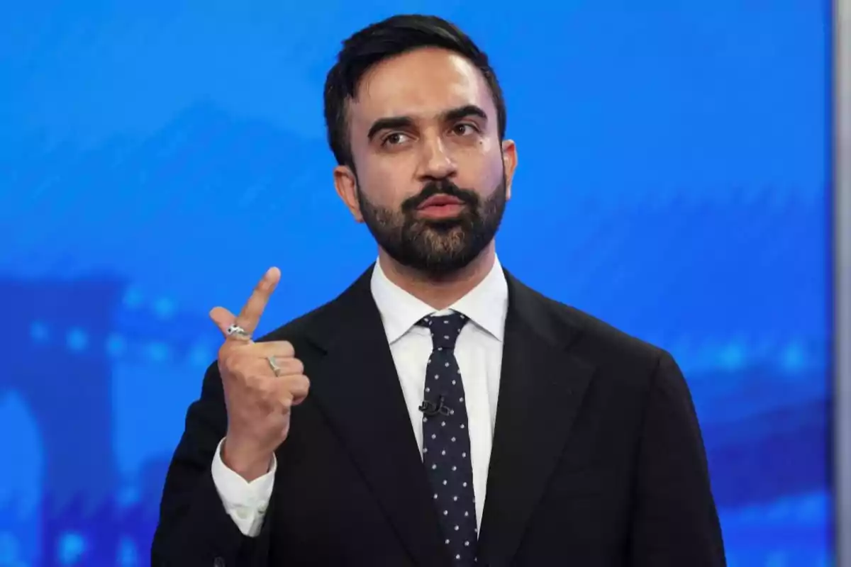 Hombre de barba y cabello oscuro vestido con traje y corbata azul con puntos blancos, hablando y gesticulando con la mano frente a un fondo azul