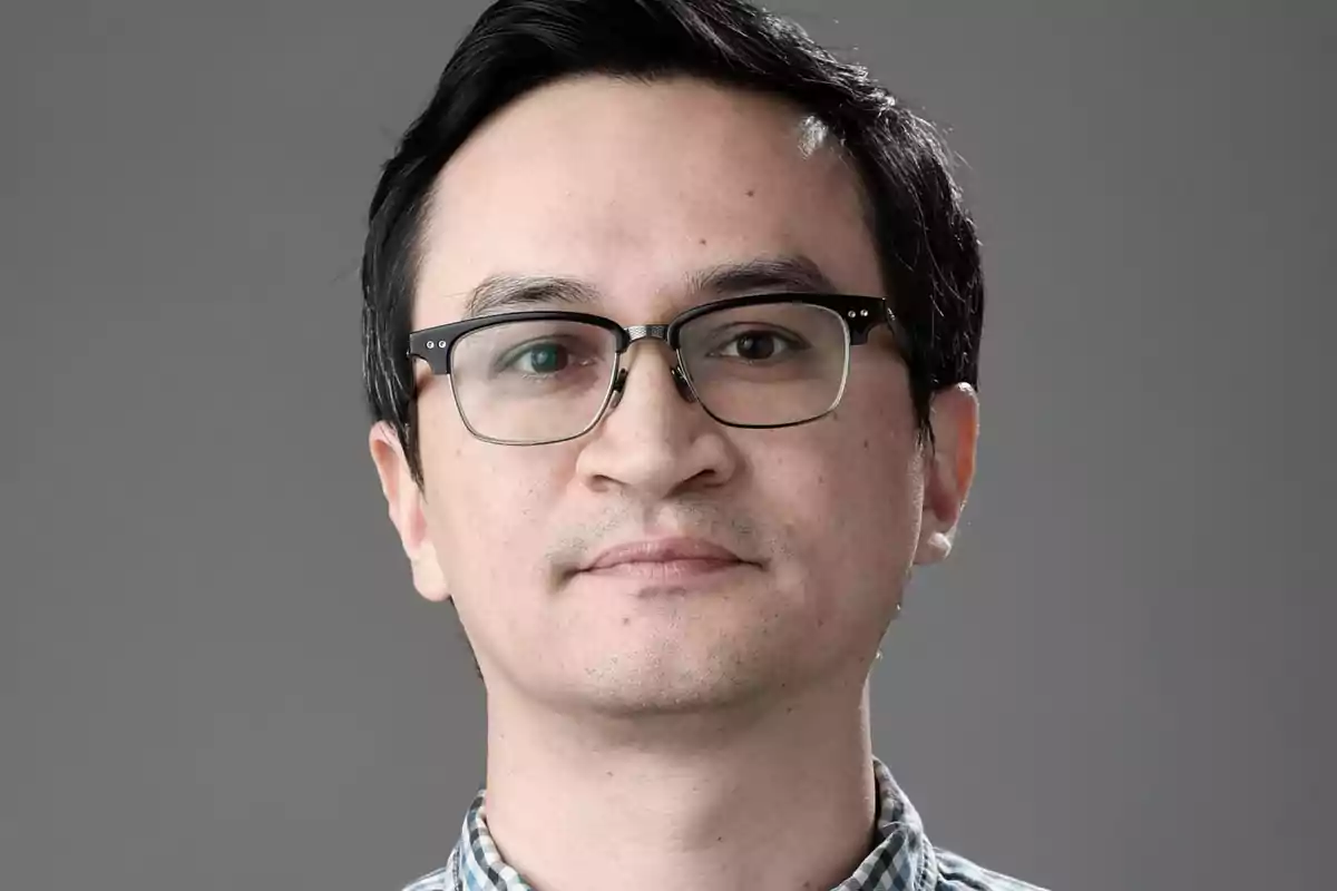 Hombre joven con gafas y camisa de cuadros mirando al frente con expresión neutral y fondo gris