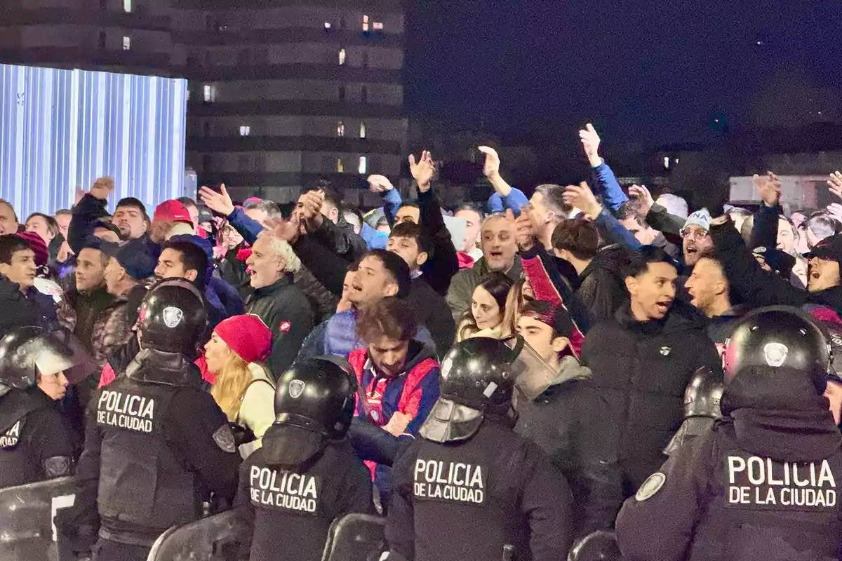 A group of people is protesting behind a police line at night while several officers wearing helmets and vests observe the situation. A group of people is protesting behind a police line at night while several officers wearing helmets and vests observe the situation.