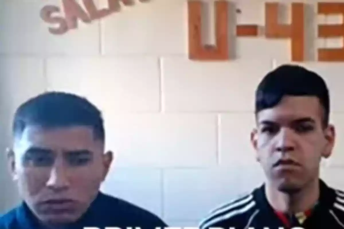 Two young men with serious expressions are sitting in front of a white wall with orange letters at the top.