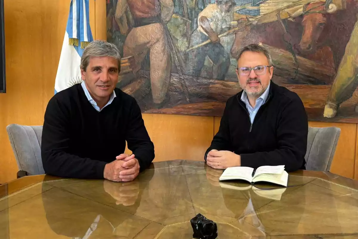 Dos hombres sentados en una mesa de vidrio con un cuaderno abierto, al fondo una bandera argentina y un mural colorido
