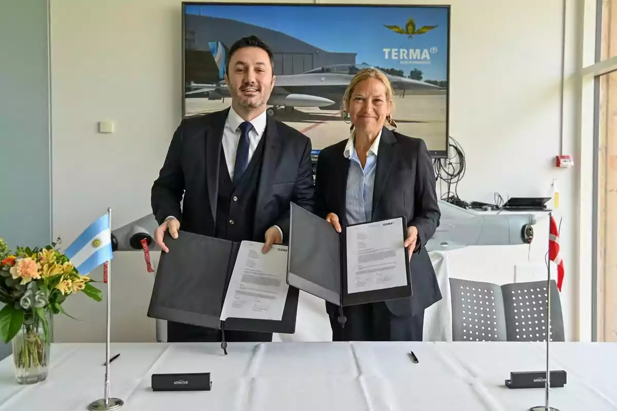 Dos personas vestidas de traje sonríen mientras muestran documentos firmados en una mesa con banderas de Argentina y Dinamarca, flores y un fondo con la imagen de un avión y el logo de Terma. Dos personas vestidas de traje sonríen mientras muestran documentos firmados en una mesa con banderas de Argentina y Dinamarca, flores y un fondo con la imagen de un avión y el logo de Terma.
