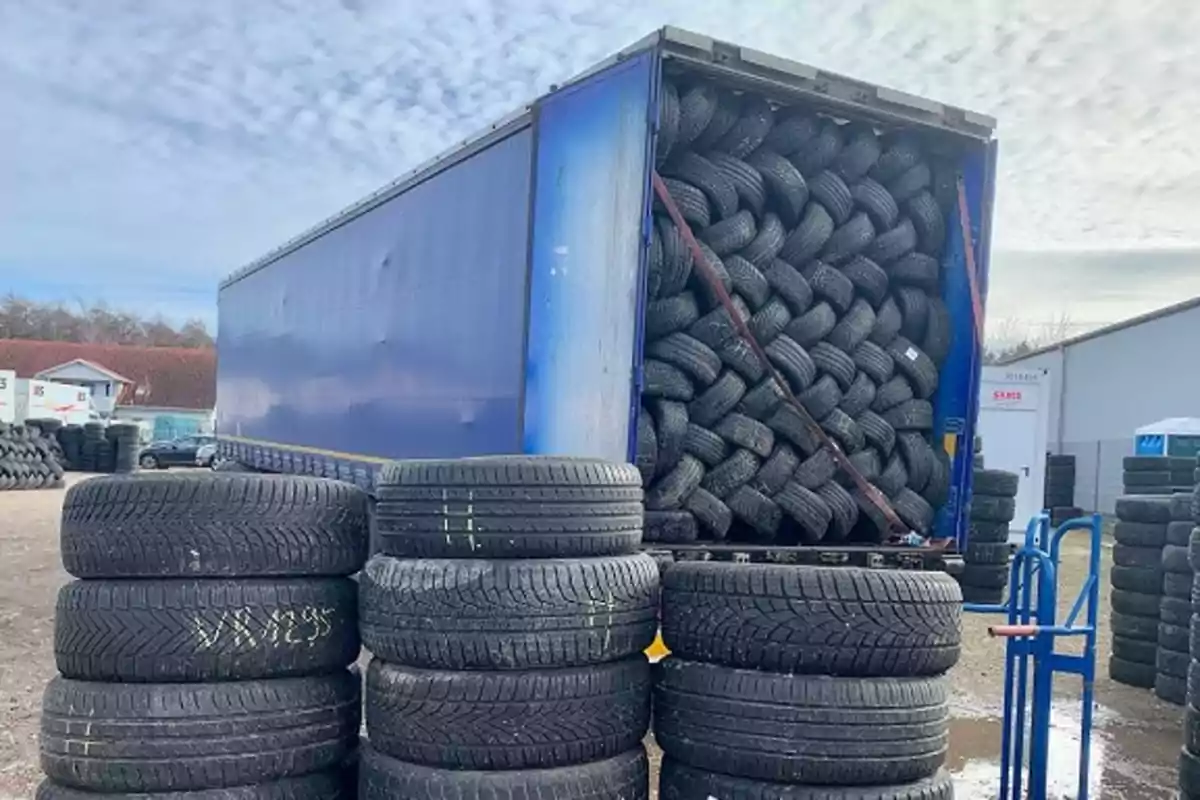 Un tráiler azul con la puerta trasera abierta está completamente lleno de llantas apiladas y en el exterior hay varias pilas de neumáticos en un patio industrial