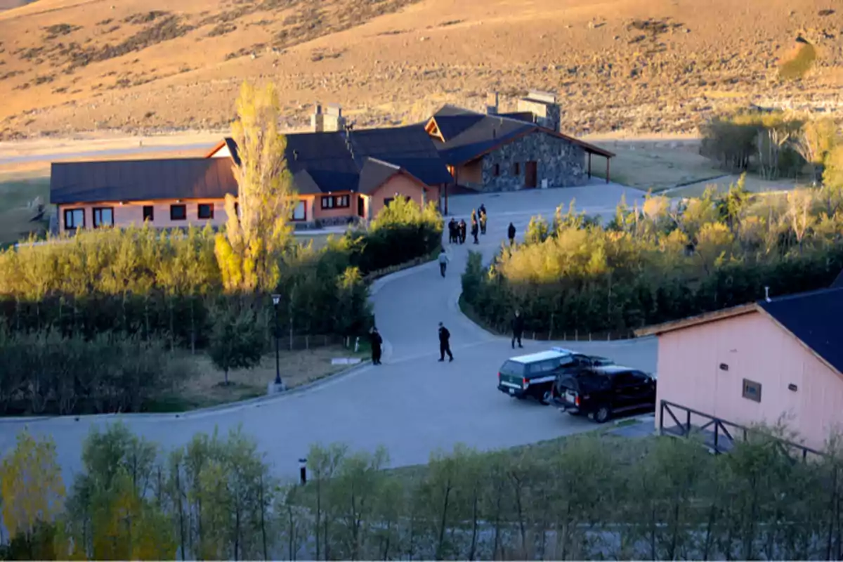 Vista aérea de una casa grande en una zona rural rodeada de árboles y colinas con varias personas caminando y algunos autos estacionados en el camino de entrada Vista aérea de una casa grande en una zona rural rodeada de árboles y colinas con varias personas caminando y algunos autos estacionados en el camino de entrada
