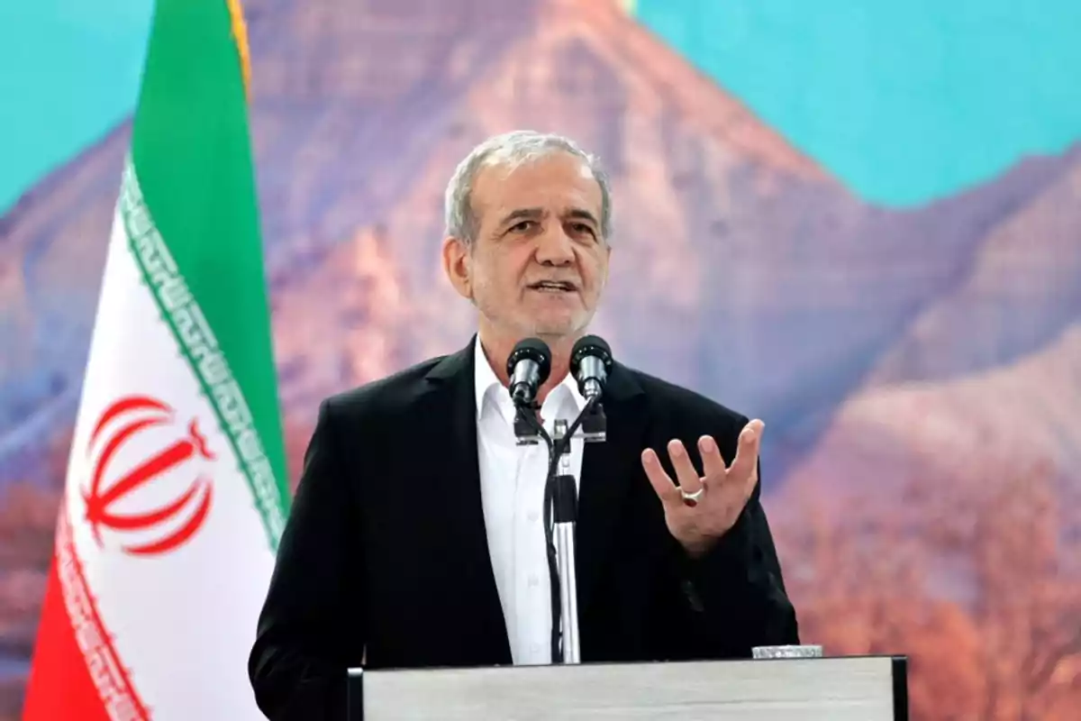 Hombre de cabello canoso hablando en un podio con micrófonos, al fondo una bandera de Irán y un paisaje montañoso.