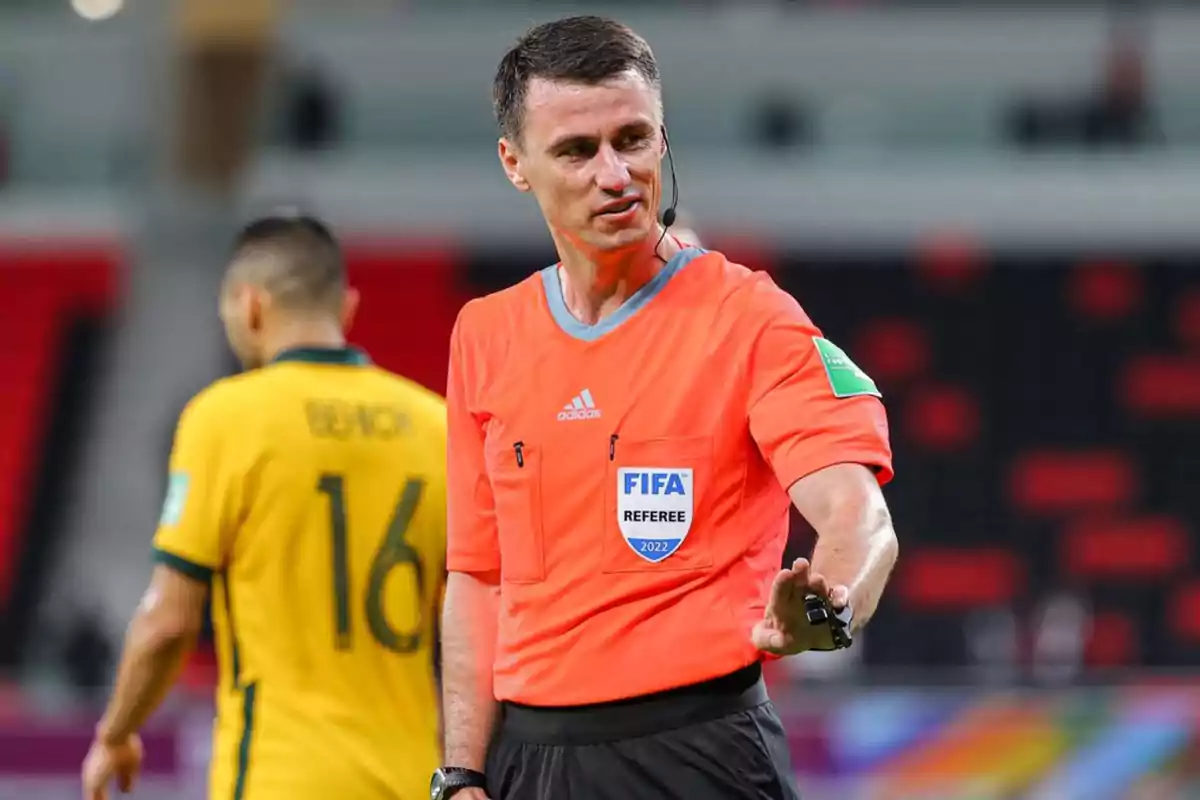Árbitro de fútbol con uniforme naranja señalando con la mano durante un partido, al fondo se ve un jugador con camiseta amarilla y el número 16. Árbitro de fútbol con uniforme naranja señalando con la mano durante un partido, al fondo se ve un jugador con camiseta amarilla y el número 16.