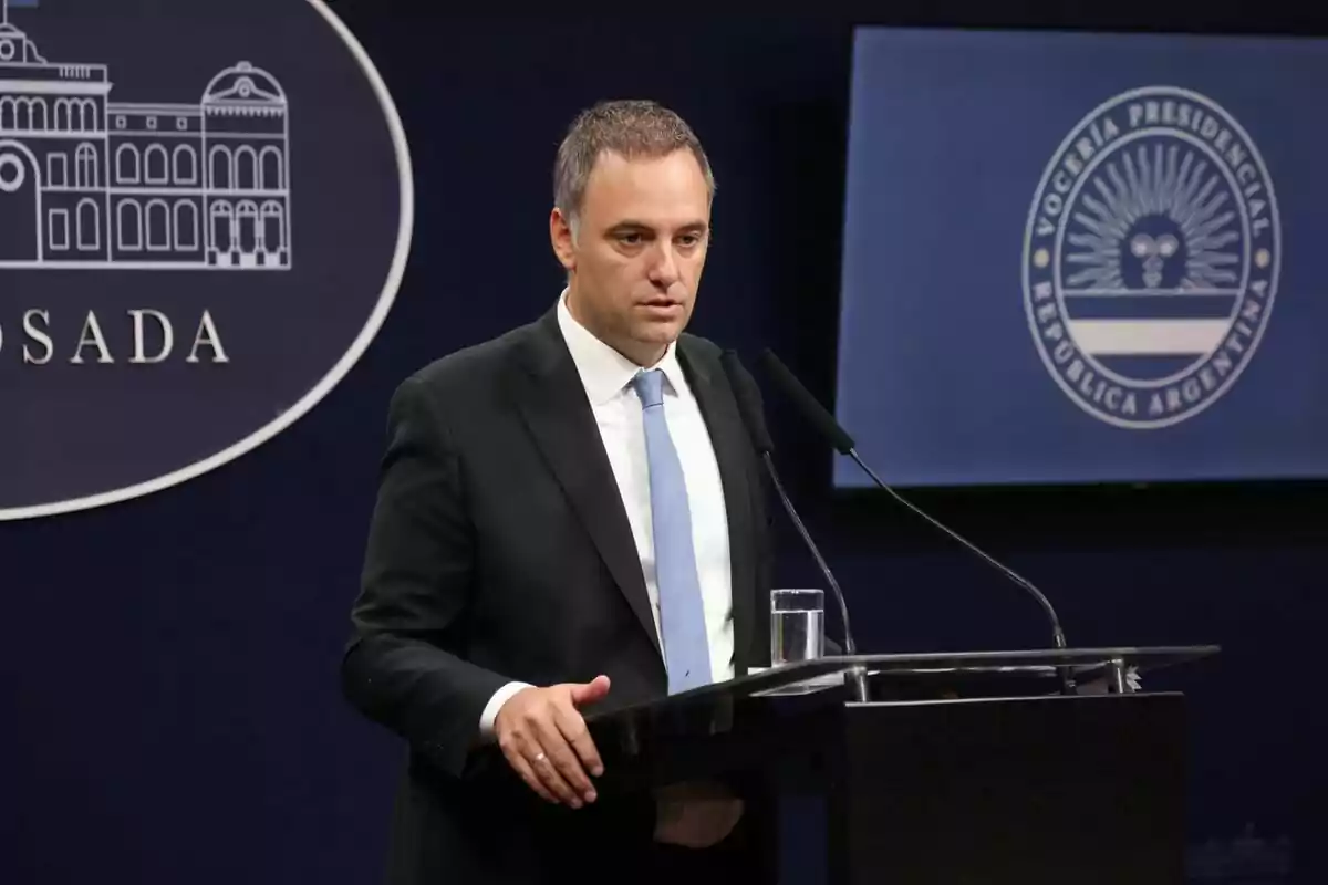 A man in a suit and light blue tie speaks at a press conference at a podium with microphones and a glass of water; in the background, official logos of the Argentine government can be seen.