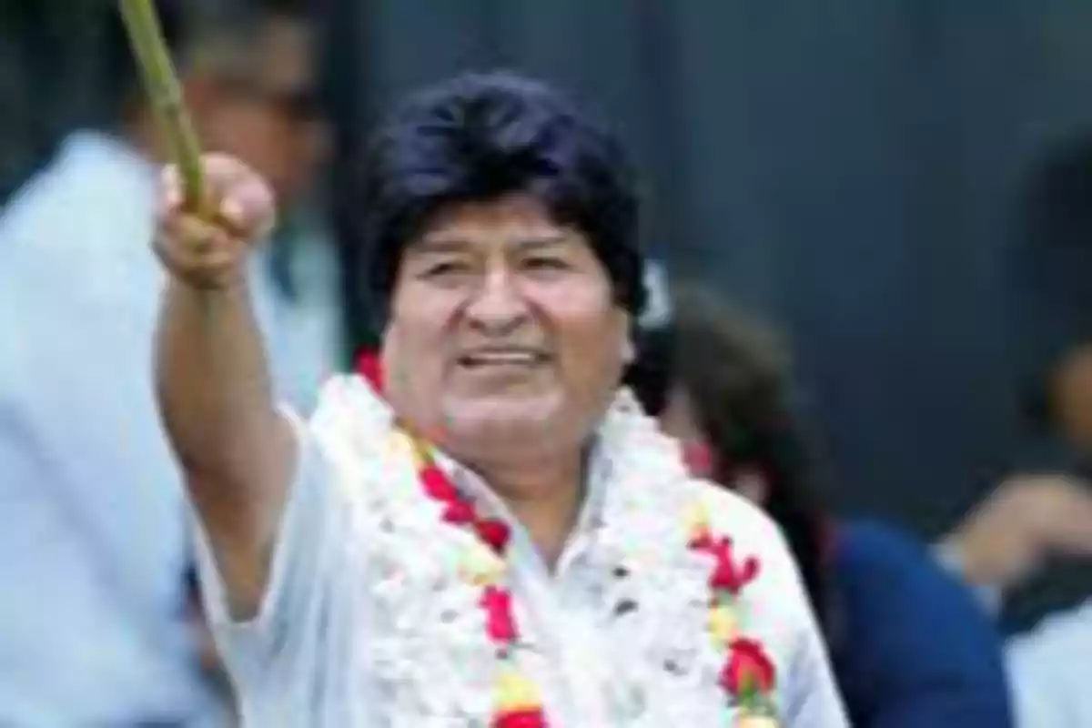 Hombre de cabello oscuro con guirnalda de flores blancas y rojas levantando un brazo en un evento público Hombre de cabello oscuro con guirnalda de flores blancas y rojas levantando un brazo en un evento público