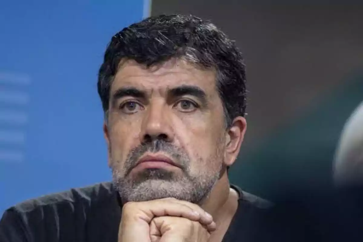 Hombre de cabello oscuro y barba con expresión seria apoyando el mentón sobre su mano Hombre de cabello oscuro y barba con expresión seria apoyando el mentón sobre su mano