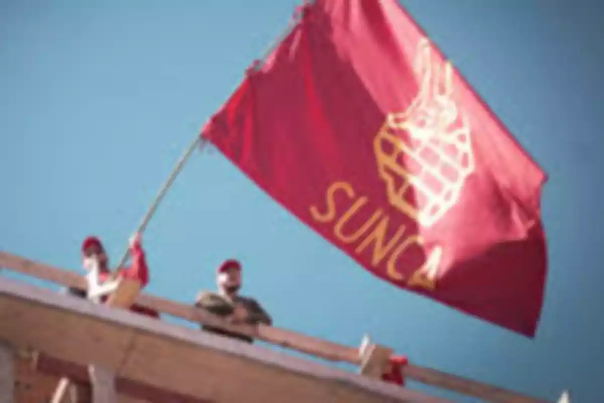 People wearing red caps hold a red flag with the SUNCA logo and name on a wooden structure under a clear sky. People wearing red caps hold a red flag with the SUNCA logo and name on a wooden structure under a clear sky.