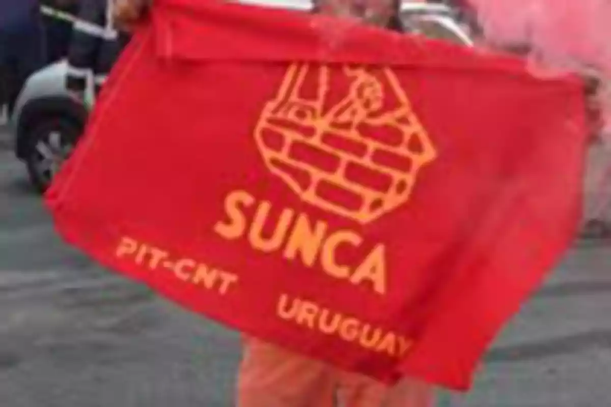 Persona sosteniendo una bandera roja del sindicato SUNCA de Uruguay con letras y un logo en amarillo Persona sosteniendo una bandera roja del sindicato SUNCA de Uruguay con letras y un logo en amarillo