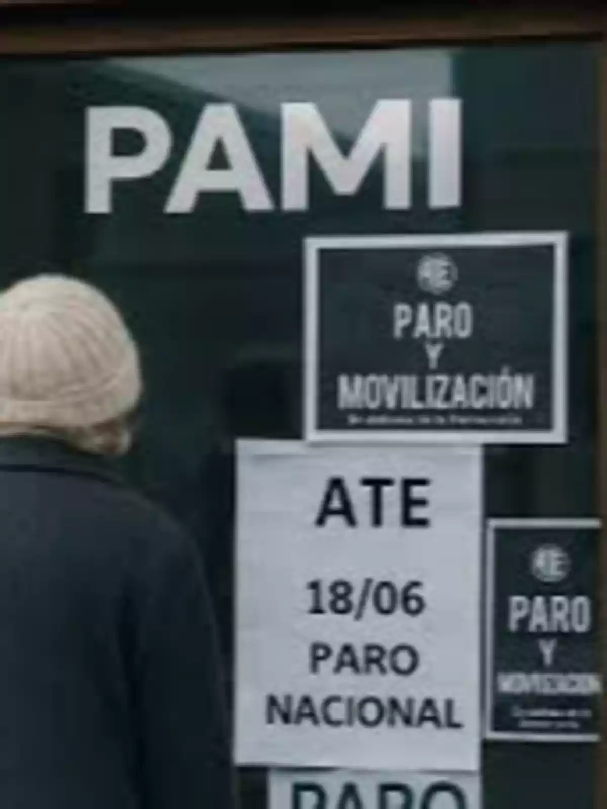 Persona de espaldas con gorro de lana frente a una puerta de PAMI con carteles de paro y movilización y un aviso de paro nacional convocado por ATE el 18 de junio