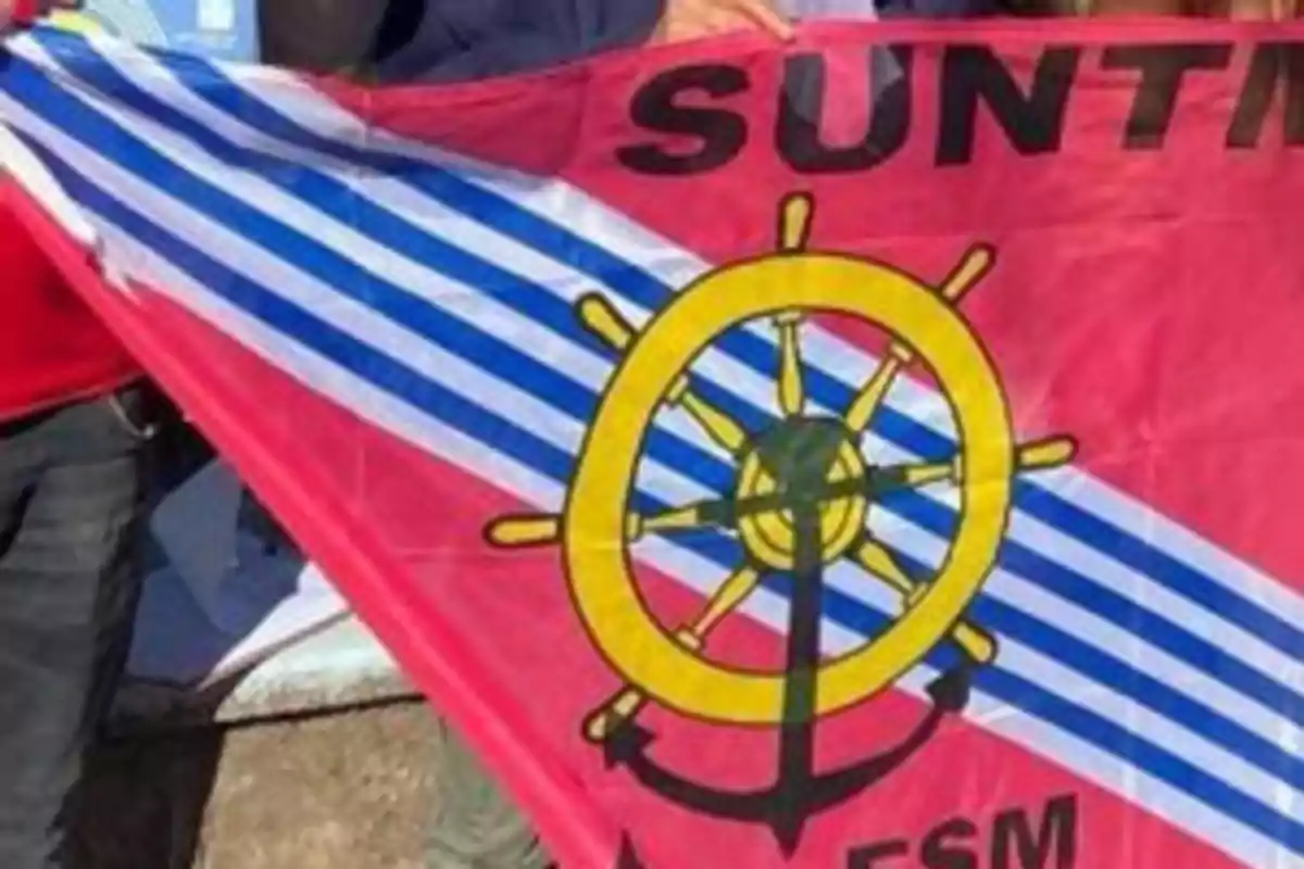 Bandera roja con franjas diagonales azules y blancas, un timón amarillo y un ancla negra en el centro, con letras negras en la parte superior.